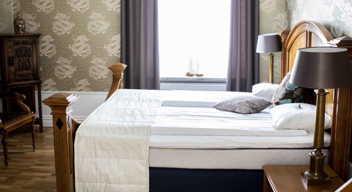 Hotellrum med modernt bäddad dubbelsäng i förgrunden och ett fönster med lila gardiner i bakgrunden.