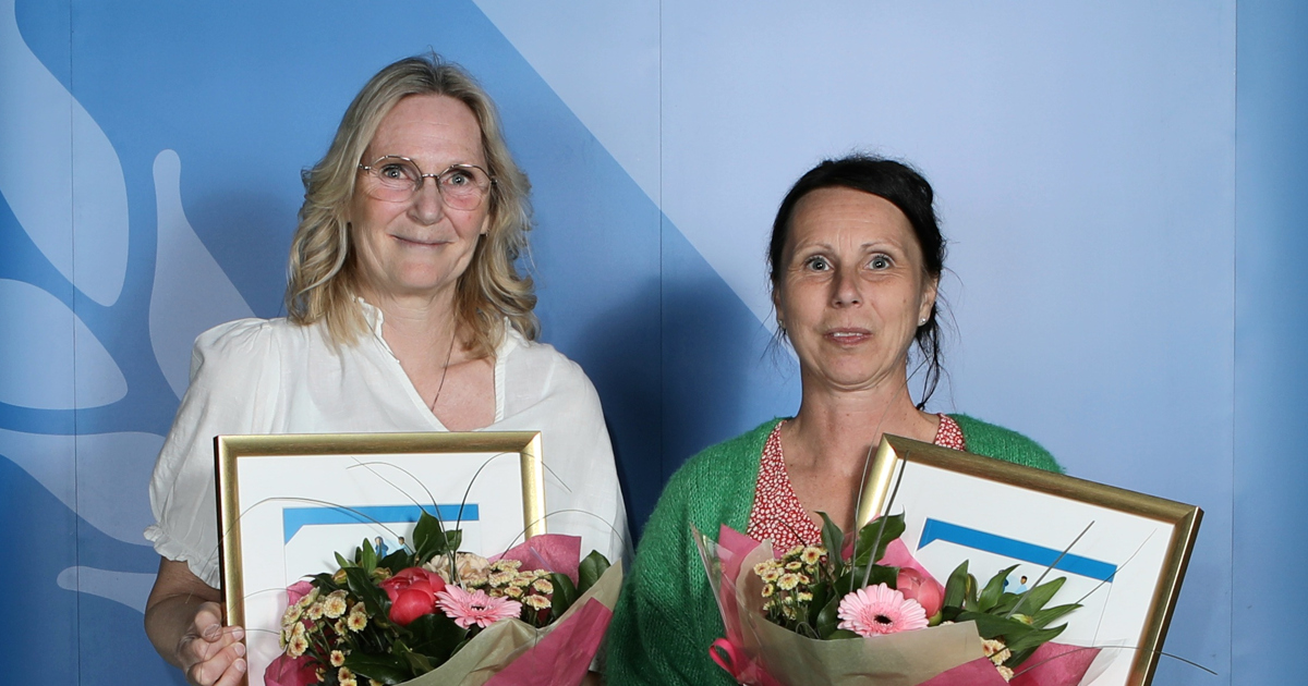 Två kvinnor står framför en kuliss och håller i blommor.