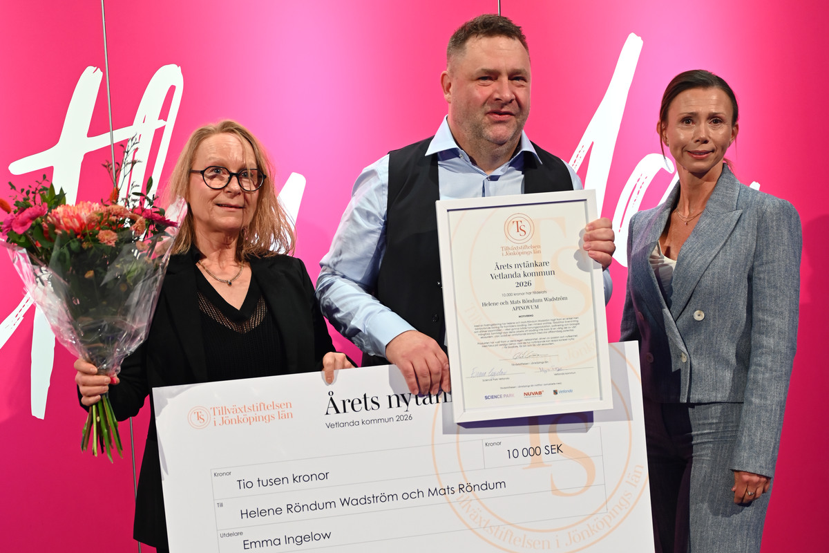 En man och en kvinna håller i en stor check och i händerna håller de ett diplom och en blombukett. Bredvid står en kvinna som har delat ut priset till dem.