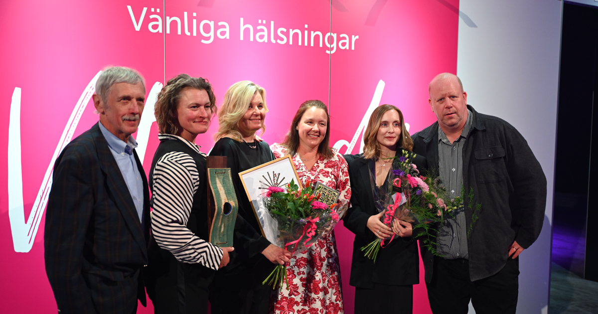 Två män och fyra kvinnor håller i statyett, diplom och blombukett framför en fotovägg.