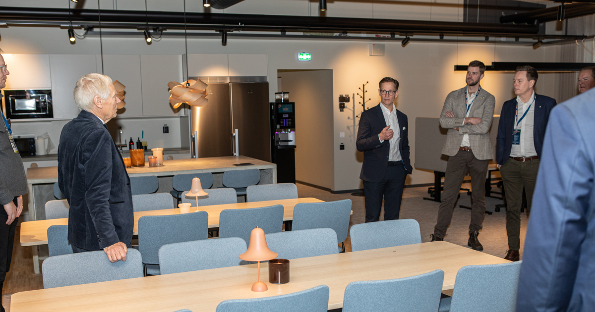 En samling människor står i en lokal med kök och flera bord.