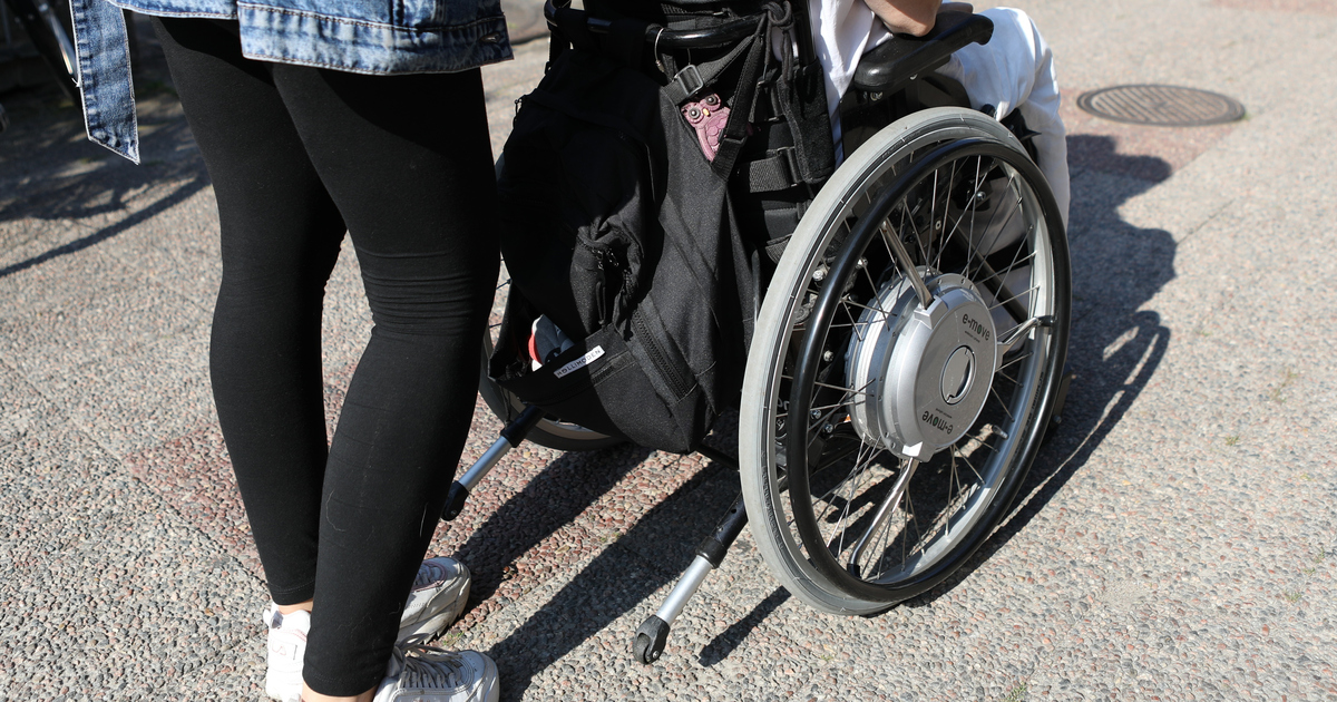 Närbild på person i rullstol med assistent bakom sig.