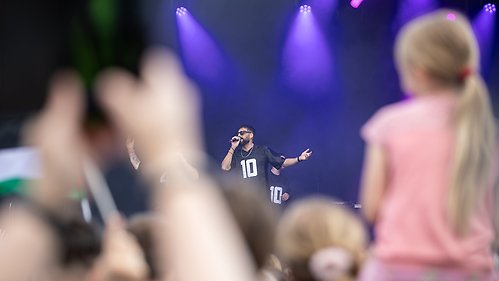 I förgrunden syns suddiga människor i publiken, längst bort i bild syns en sångare stå med en mikrofon på en scen.